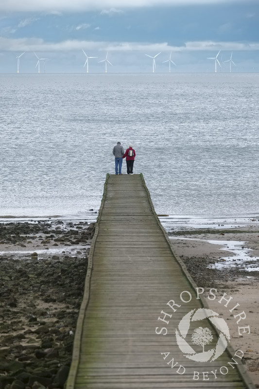 An elderly couple look out across the sea towards Gwynt y Mor wind farm from a jetty at Llandudno, Conwy, Wales.