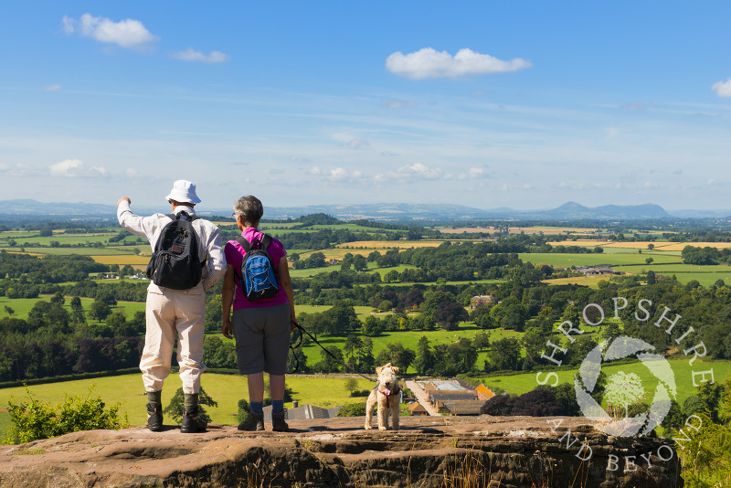 A pair of walkers, accompanied by their dog, look out over the north Shropshire countryside from Grinshill Hill, Shropshire, England