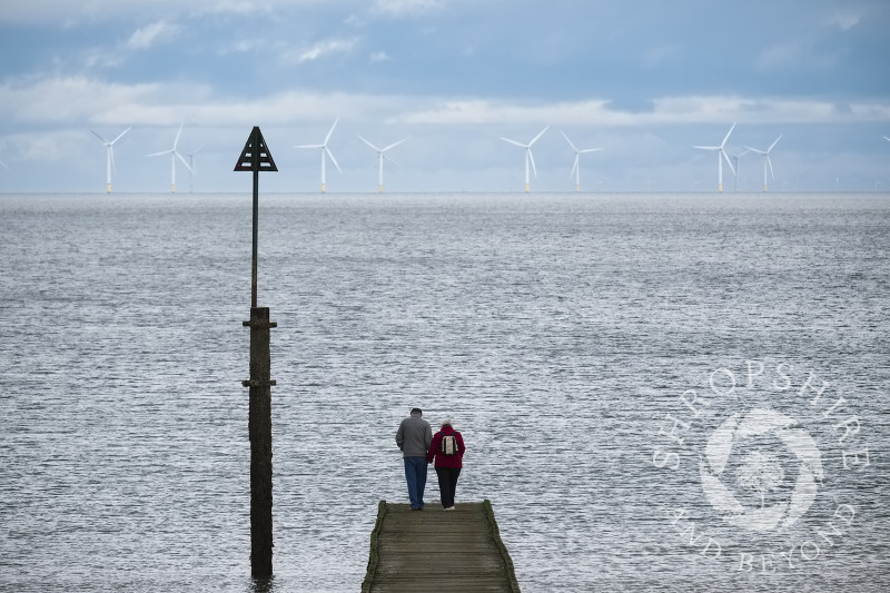 An elderly couple stand on a jetty at Llandudno, Conwy, Wales and look out across the sea towards Gwynt y Mor wind farm.