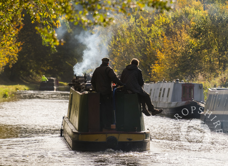 A narrowboat emits smoke from a wood burning stove amid autumn colour on the Shropshire Union Canal at Brewood, Staffordshire.
